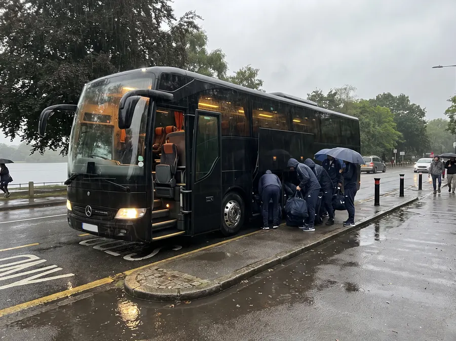 A black executive coach waiting on the road beside Ruislip Lido in steady rain, headlights on and door open, a sports team in matching tracksuits loading kit bags into the luggage bay with the lake and woodland behind them.