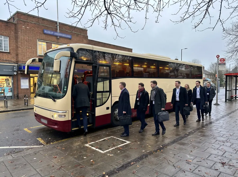 A cream and burgundy touring coach on the forecourt outside Ruislip Underground station on an overcast winter afternoon, its door open and interior lit, a corporate group in business-casual clothes with laptop bags and lanyards boarding in front of the brick station building.